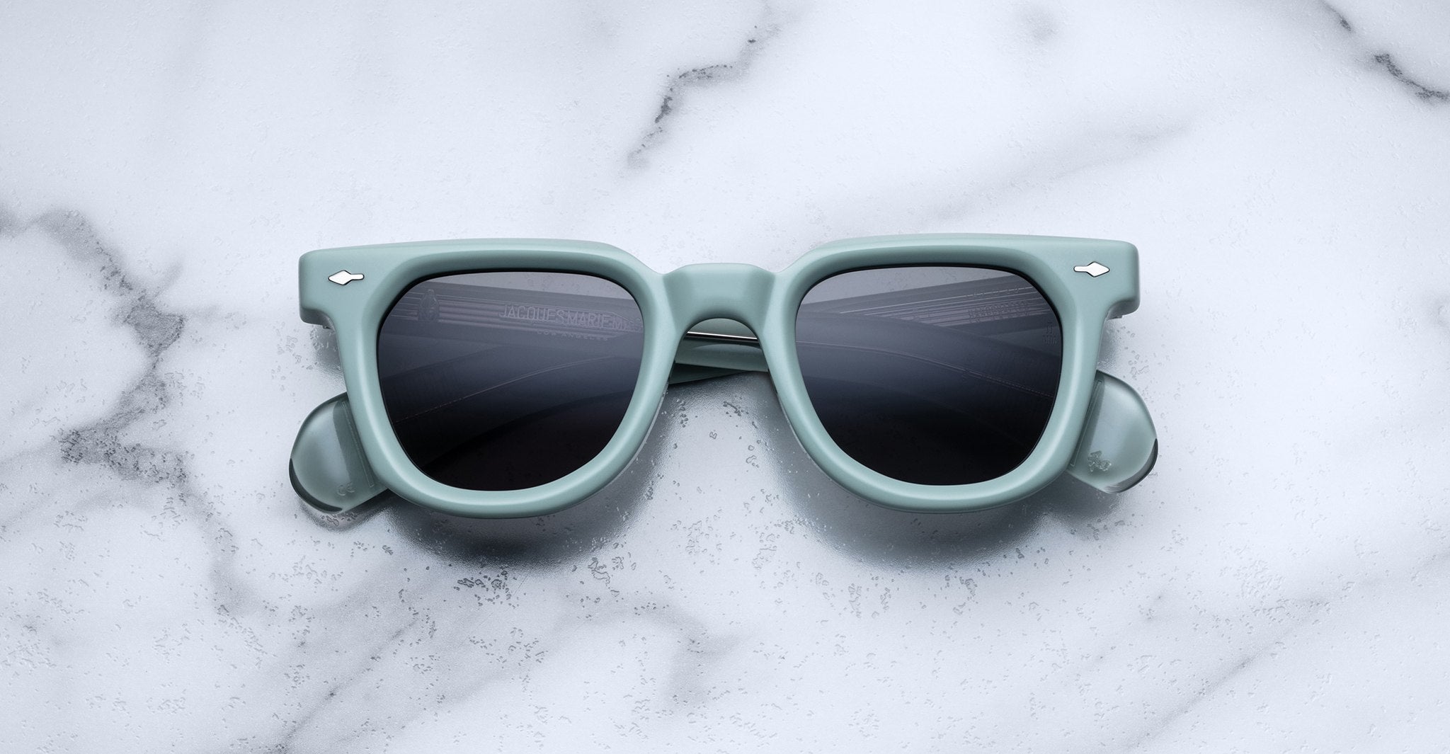 A pair of Jacques Marie Mage Vendome sunglasses in mint green with dark lenses sits atop a white marble surface.