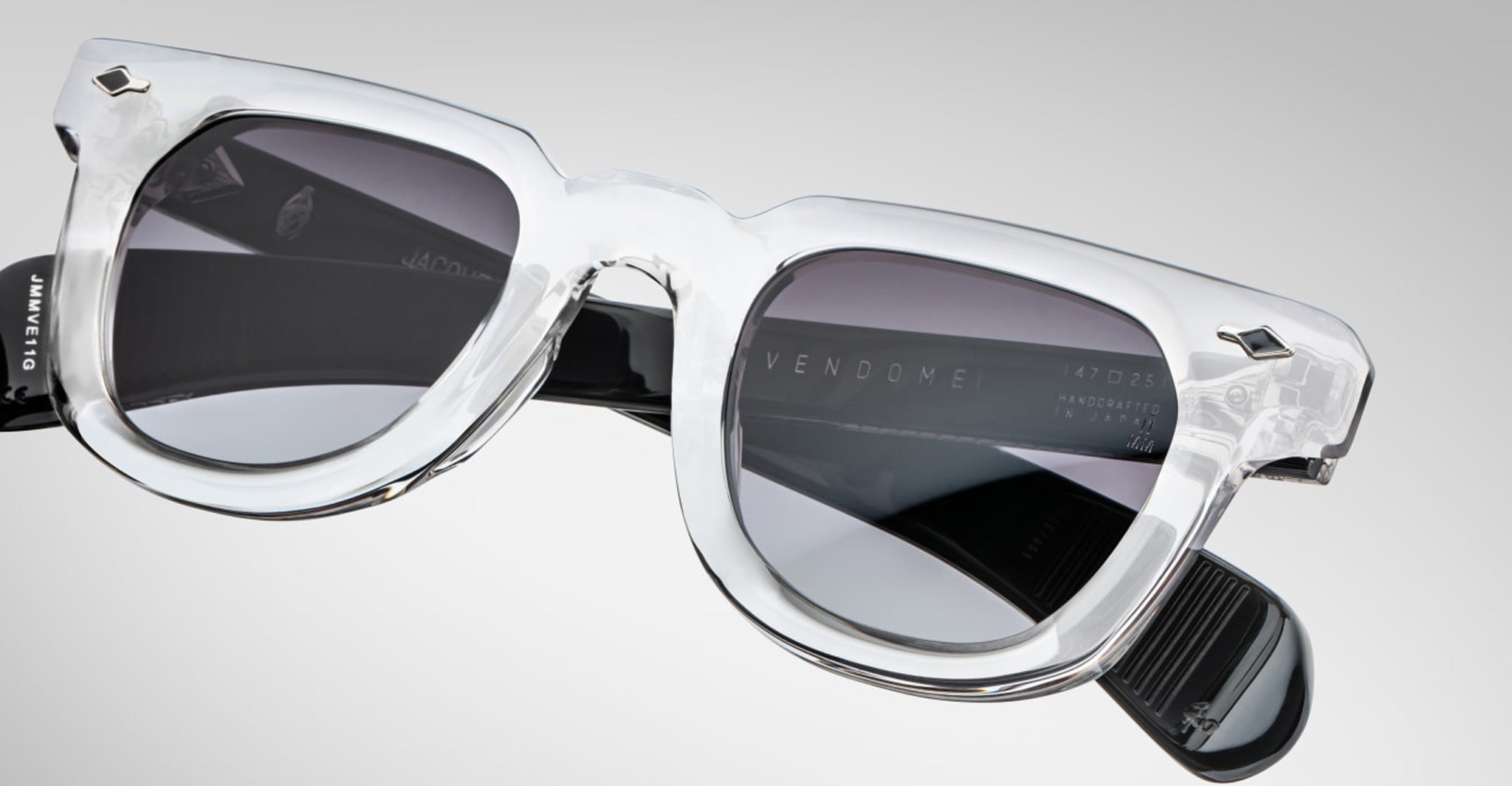 Jacques Marie Mage Vendome sunglasses featuring clear frames, black arms, and dark lenses are handcrafted in Japan and displayed on a plain light gray background.