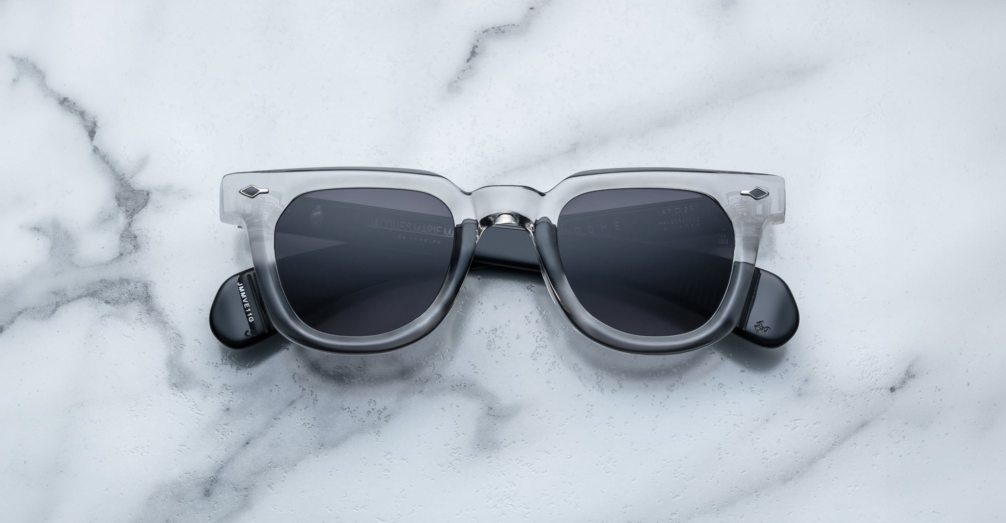 A pair of Jacques Marie Mage Vendome sunglasses by Jacques Marie Mage, featuring gray translucent frames and dark lenses, rests on a white marble surface.