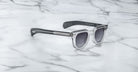 A pair of Jacques Marie Mage Vendome sunglasses by Jacques Marie Mage, featuring clear frames and dark lenses, handcrafted in Japan, elegantly displayed on a white marble surface.