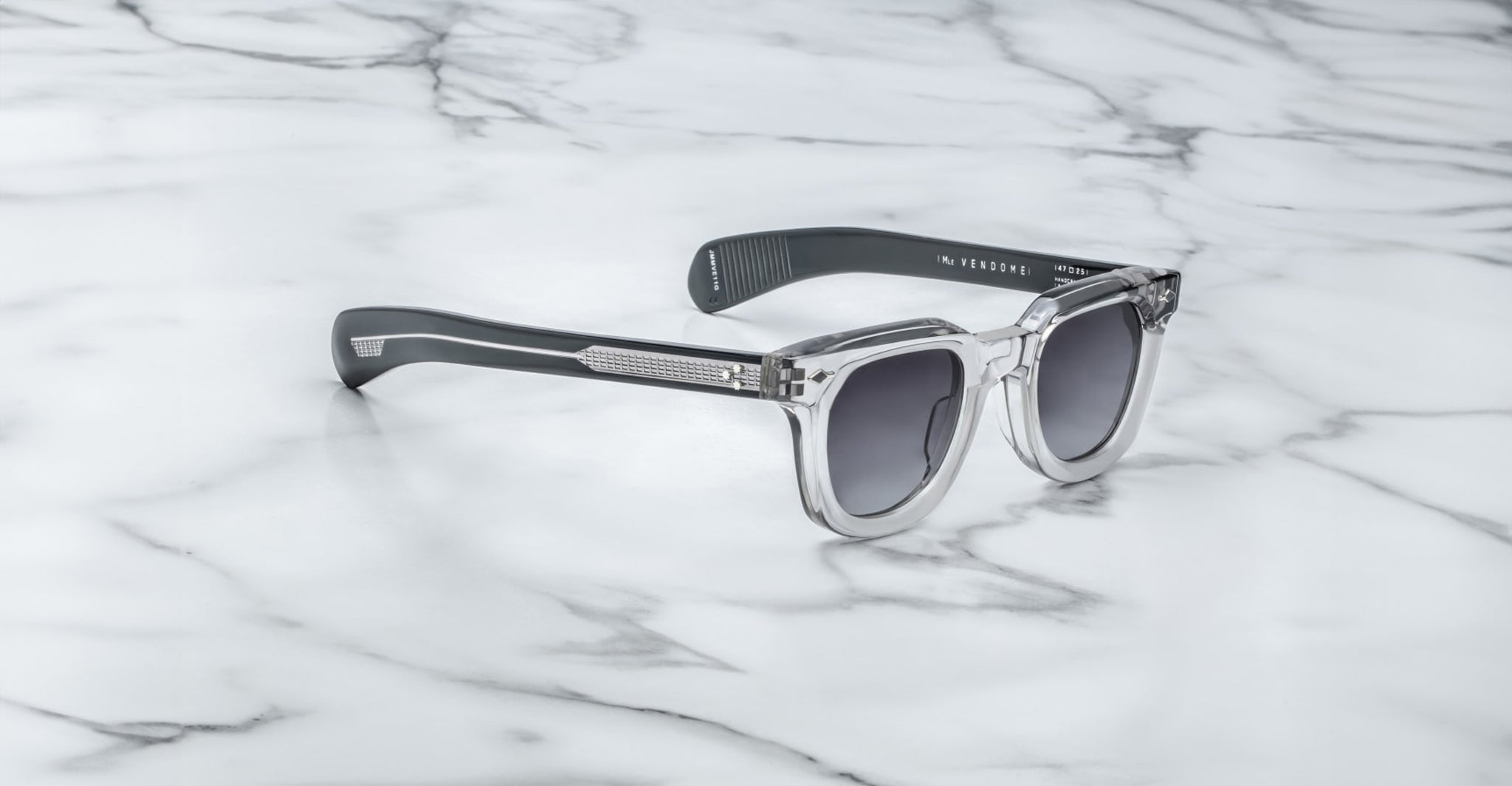 A pair of Jacques Marie Mage Vendome sunglasses by Jacques Marie Mage, featuring clear frames and dark lenses, handcrafted in Japan, elegantly displayed on a white marble surface.