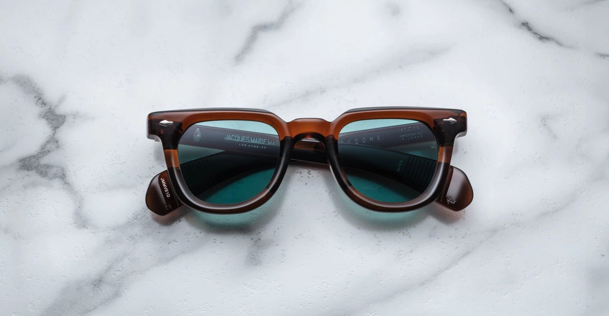A pair of Jacques Marie Mage Vendome sunglasses by Jacques Marie Mage, featuring brown frames and blue-green tinted lenses, handcrafted in Japan, displayed on a white marble surface.