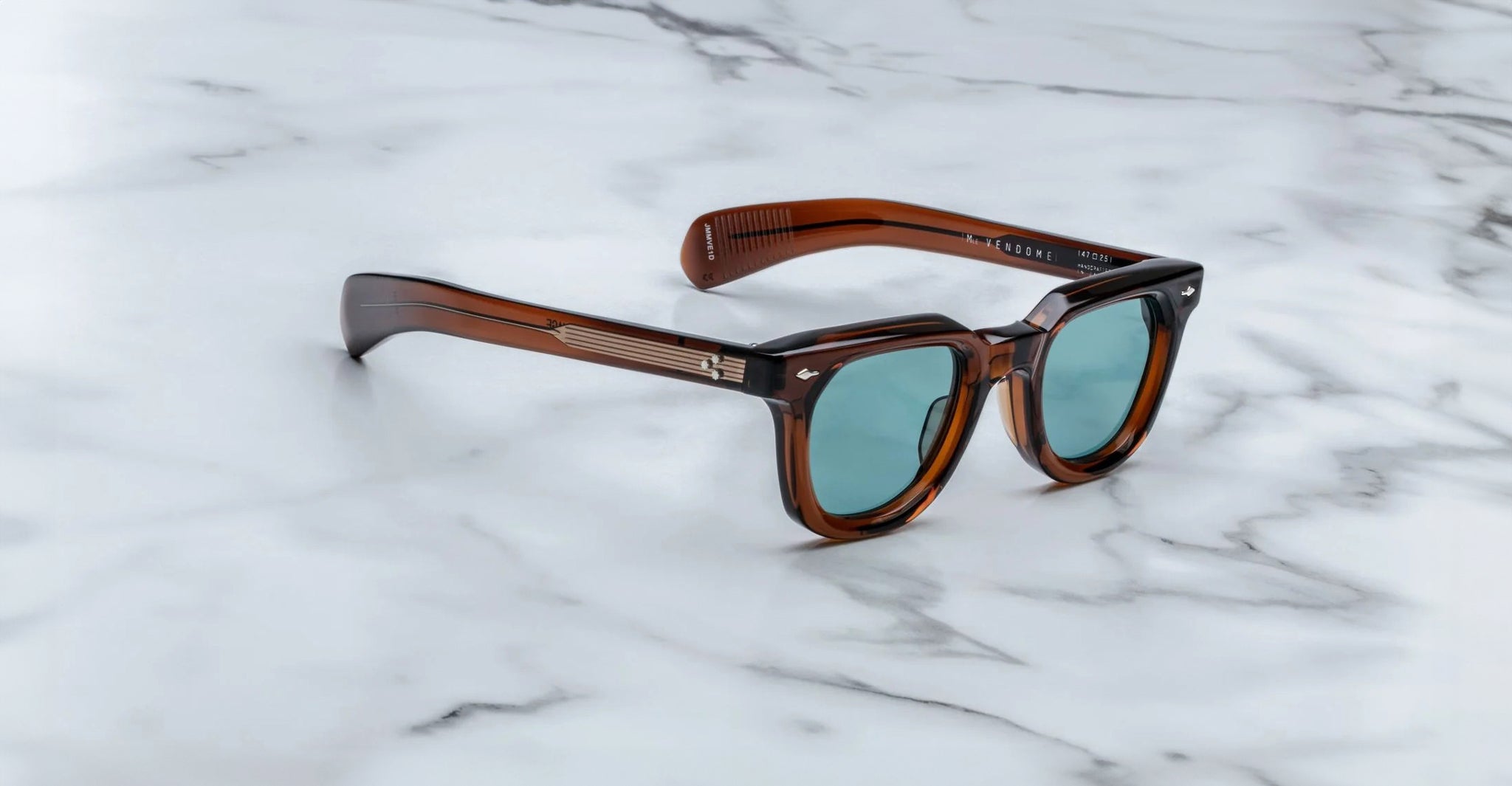 A pair of Jacques Marie Mage Vendome sunglasses by Jacques Marie Mage, featuring a brown rectangular frame and teal lenses, handcrafted in Japan, rests on a white marble surface.