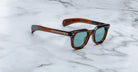 A pair of Jacques Marie Mage Vendome sunglasses by Jacques Marie Mage, featuring a brown rectangular frame and teal lenses, handcrafted in Japan, rests on a white marble surface.