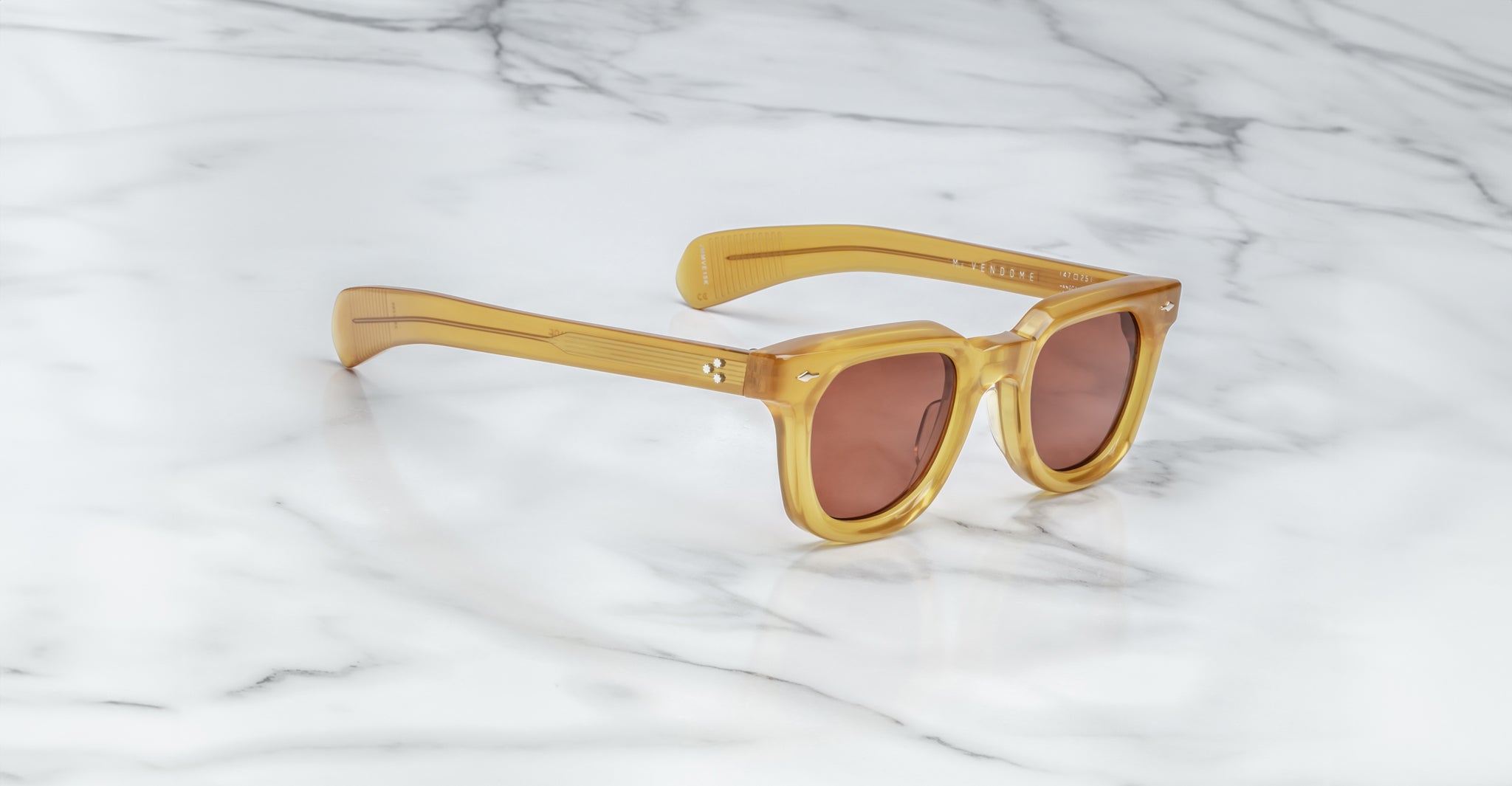 Jacques Marie Mage Vendome sunglasses with thick amber frames and brown-tinted lenses, handcrafted in Japan, are displayed on a white marble surface.