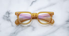 A pair of Jacques Marie Mage Vendome amber optical frames with reflective pink lenses, handcrafted in Japan, rests on a white marble surface.