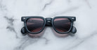 A pair of Jacques Marie Mage Vendome square sunglasses in black with dark lenses, handcrafted in Japan, rests on a white marble surface.