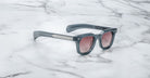 A pair of Jacques Marie Mage Vendome sunglasses by Jacques Marie Mage, featuring gray frames and pink-tinted lenses, handcrafted in Japan, is displayed on a white marble surface.