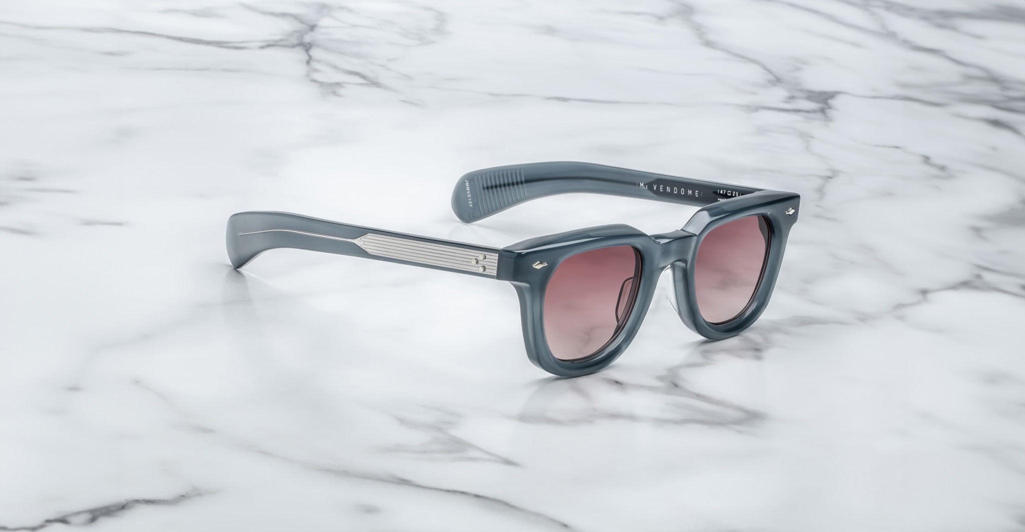 A pair of Jacques Marie Mage Vendome sunglasses by Jacques Marie Mage, featuring gray frames and pink-tinted lenses, handcrafted in Japan, is displayed on a white marble surface.