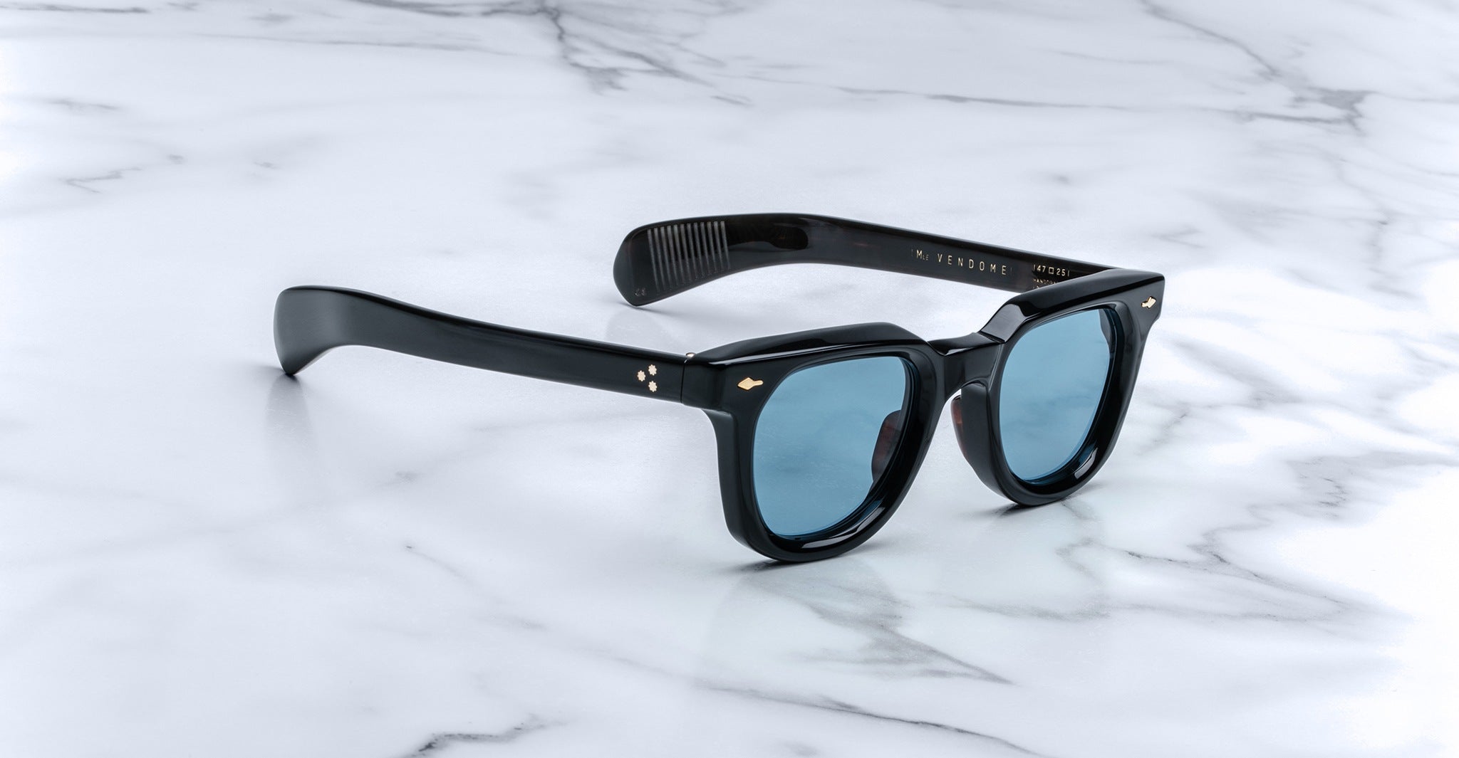 Jacques Marie Mage Vendome sunglasses by Jacques Marie Mage, featuring black frames and blue lenses, handcrafted in Japan, displayed on a white marble surface.