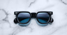 A pair of Jacques Marie Mage Vendome sunglasses by Jacques Marie Mage, featuring black frames and blue tinted lenses, handcrafted in Japan, rests on a white marble surface.