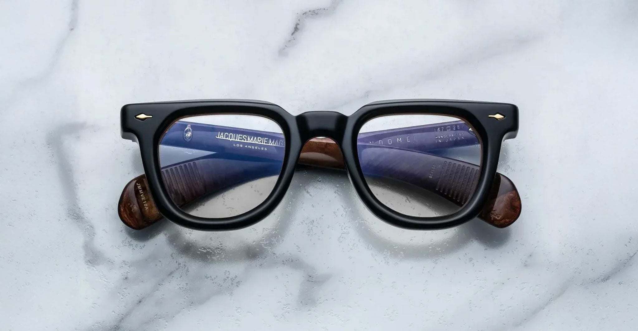 The Jacques Marie Mage Vendome by Jacques Marie Mage are black rectangular optical frames, handcrafted in Japan, featuring visible text on the inner arms and displayed on a white marble surface.