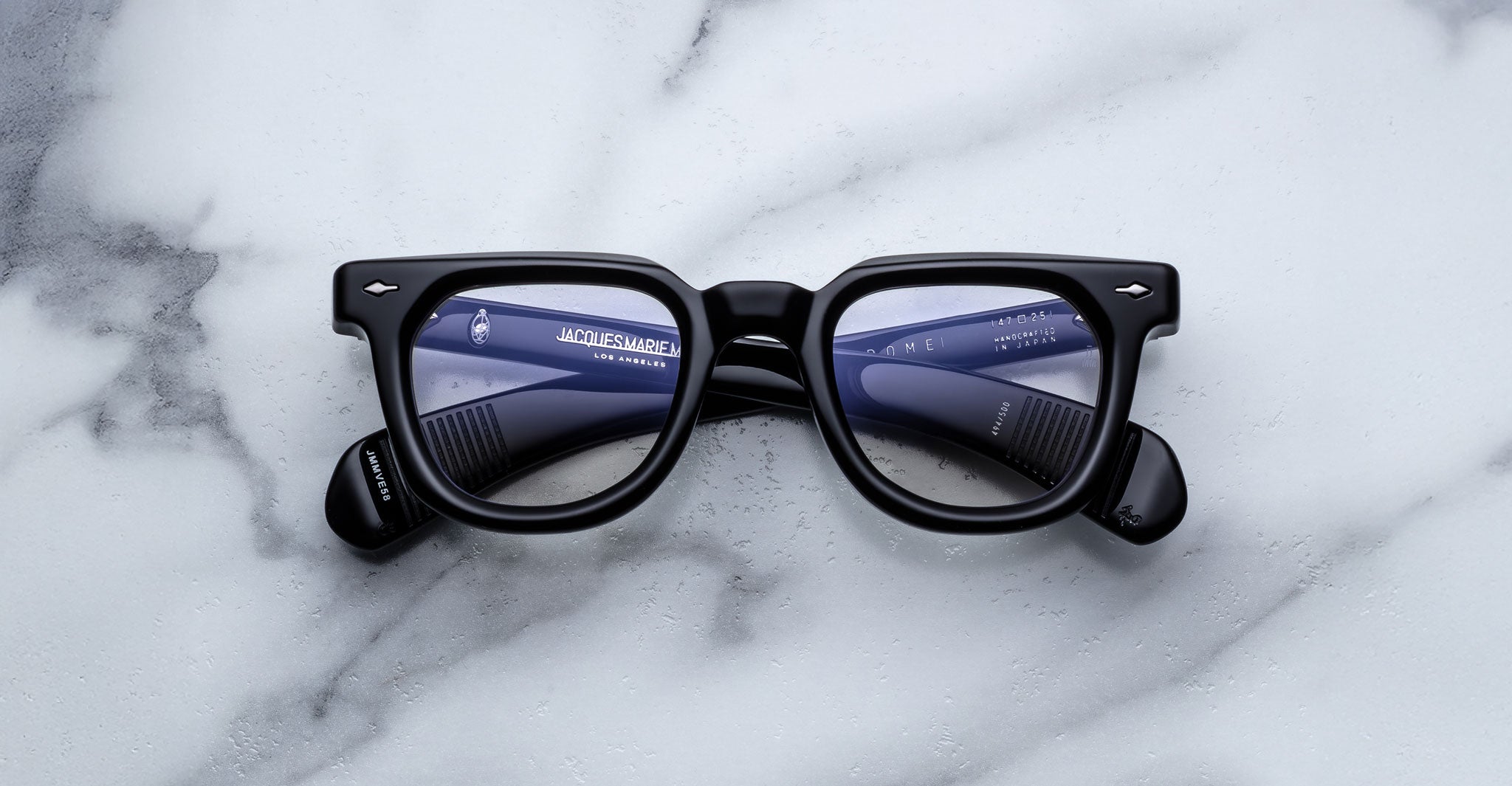 Jacques Marie Mage Vendome optical frames by Jacques Marie Mage, handcrafted in Japan, rest on white marble; their lenses reflect text and a hair comb placed behind the glasses.