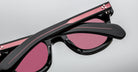 Close-up of folded Jacques Marie Mage Vendome sunglasses in black and pink with pink-tinted lenses, handcrafted in Japan, shown on a light background.