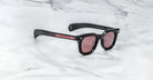 A pair of Jacques Marie Mage Vendome sunglasses by Jacques Marie Mage in black with pink-tinted lenses and red arm accents, handcrafted in Japan, displayed on a white marble surface.
