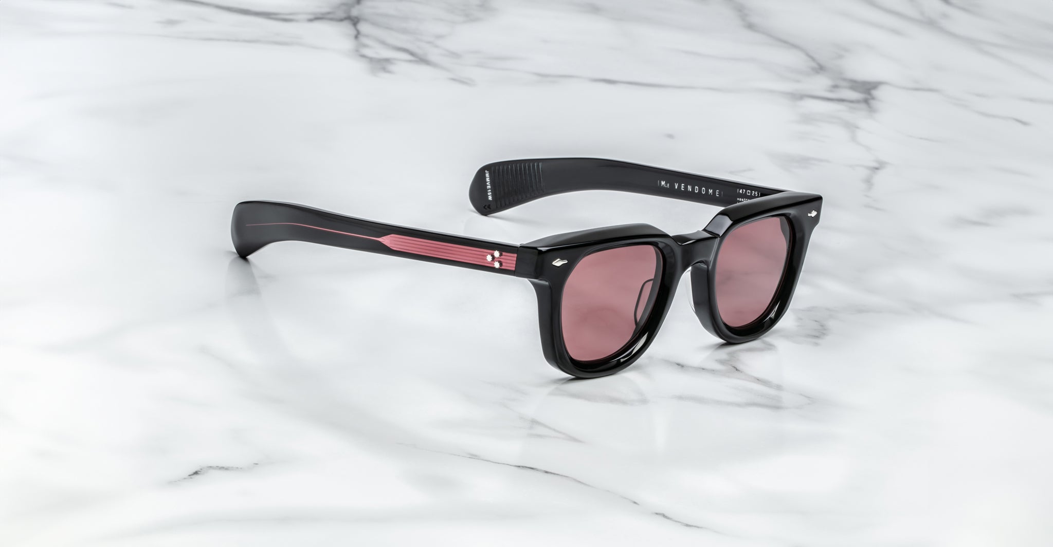 A pair of Jacques Marie Mage Vendome sunglasses by Jacques Marie Mage in black with pink-tinted lenses and red arm accents, handcrafted in Japan, displayed on a white marble surface.
