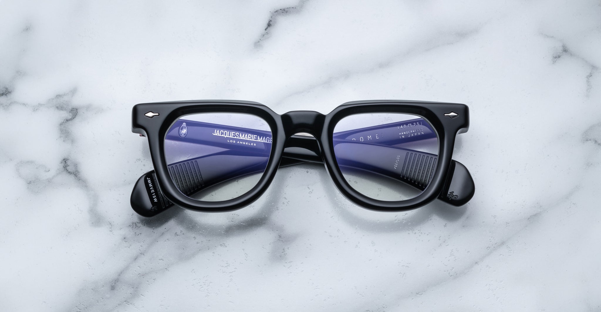 A pair of Jacques Marie Mage Vendome black optical frames by Jacques Marie Mage, handcrafted in Japan, rest on white marble with the reflection of two blue combs visible in the lenses.