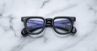 A pair of Jacques Marie Mage Vendome black optical frames by Jacques Marie Mage, handcrafted in Japan, rest on white marble with the reflection of two blue combs visible in the lenses.