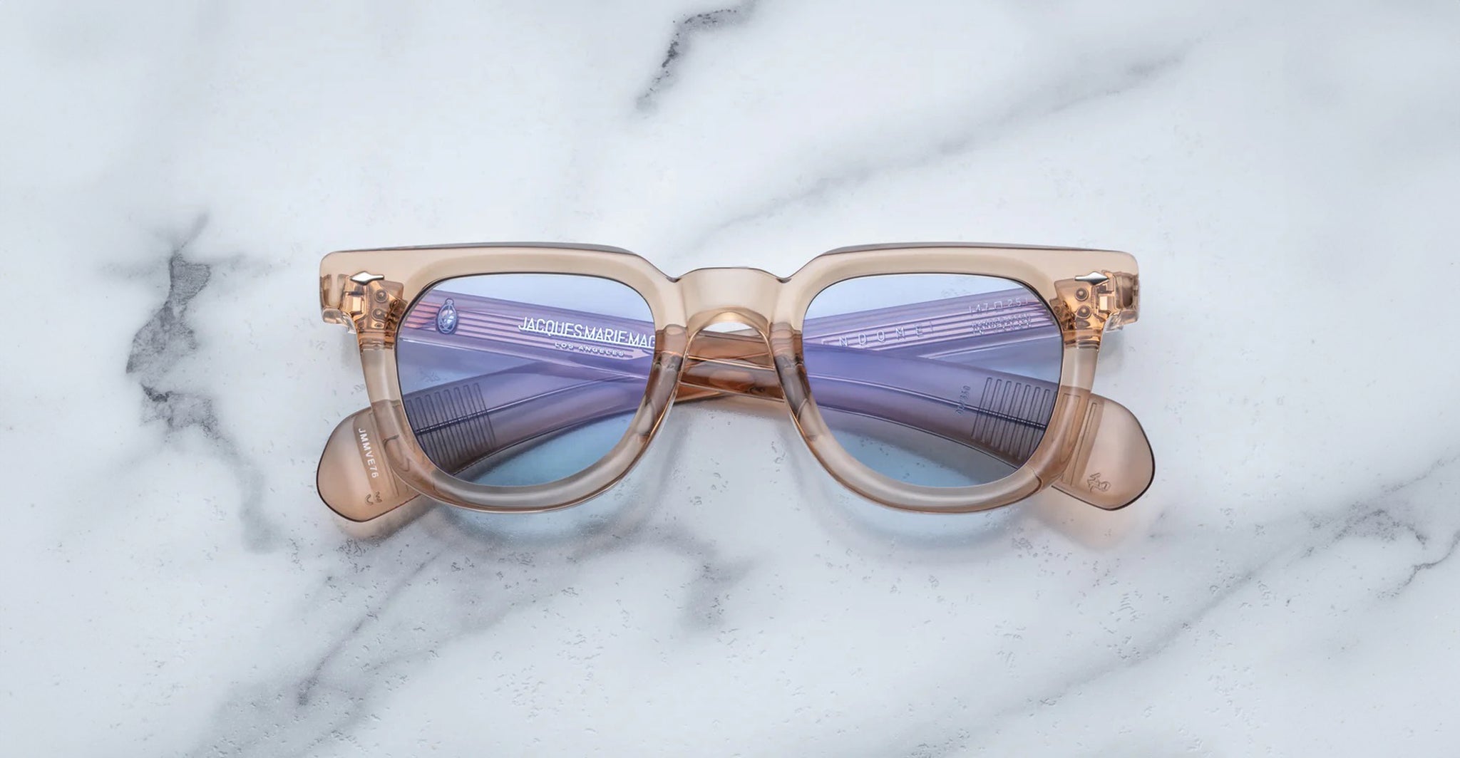 Jacques Marie Mage Vendome sunglasses by Jacques Marie Mage, featuring beige translucent frames and purple-tinted lenses inspired by Japanese craftsmanship, rest elegantly on a white marble surface.