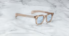 Jacques Marie Mage Vendome optical frames in translucent beige with light blue lenses rest on a white marble surface with gray veining, beautifully handcrafted in Japan.