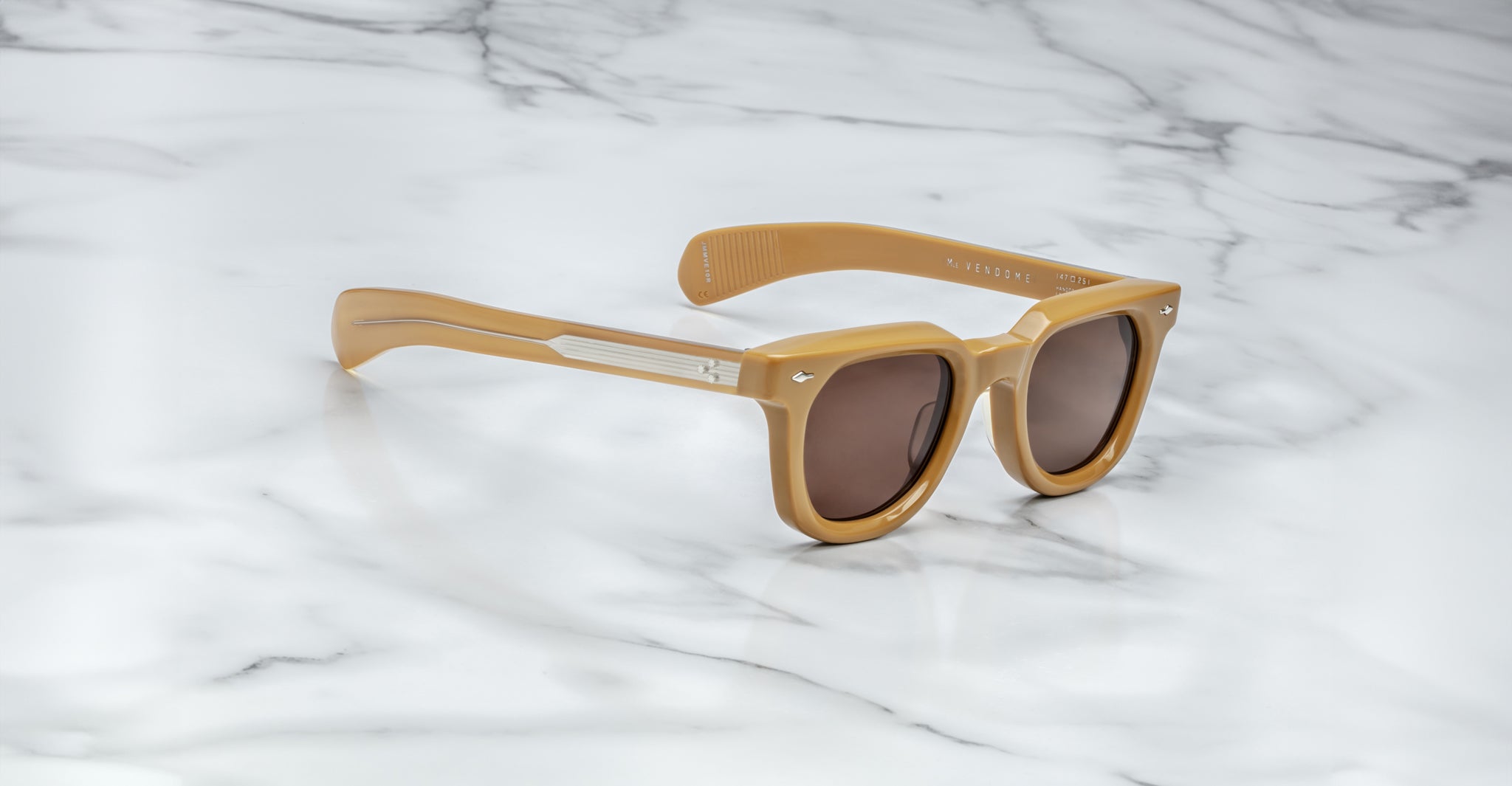 Tan plastic Jacques Marie Mage Vendome sunglasses with dark lenses and thick frames are placed on a white marble surface.