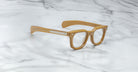 A pair of Jacques Marie Mage Vendome optical frames by Jacques Marie Mage, featuring thick tan rims and clear lenses, handcrafted in Japan, rests on a white marble surface.