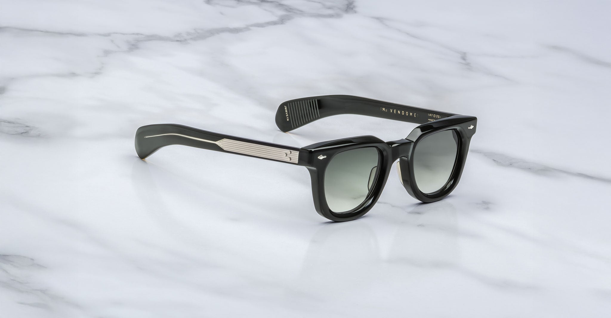 The Jacques Marie Mage Vendome sunglasses by Jacques Marie Mage, featuring thick frames and dark gradient lenses handcrafted in Japan, are elegantly displayed on a white marble surface.