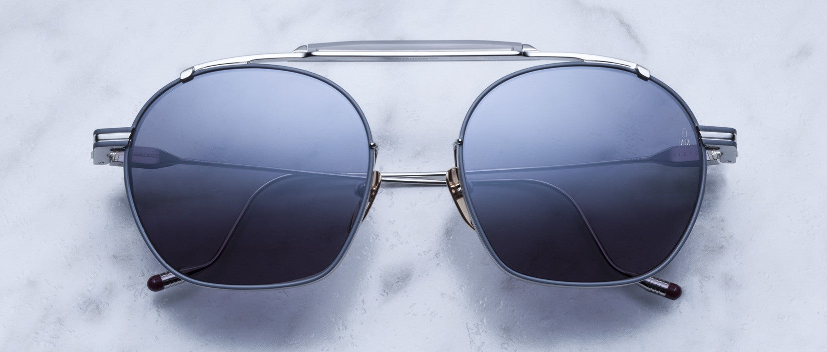 A pair of aviator-style sunglasses with dark lenses and a silver metal frame, displayed on a light, marble-textured surface.