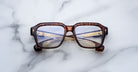 Jacques Marie Mage Vivienne square eyeglasses in brown tortoiseshell with gold decorative temples, folded and resting on a white marble surface.