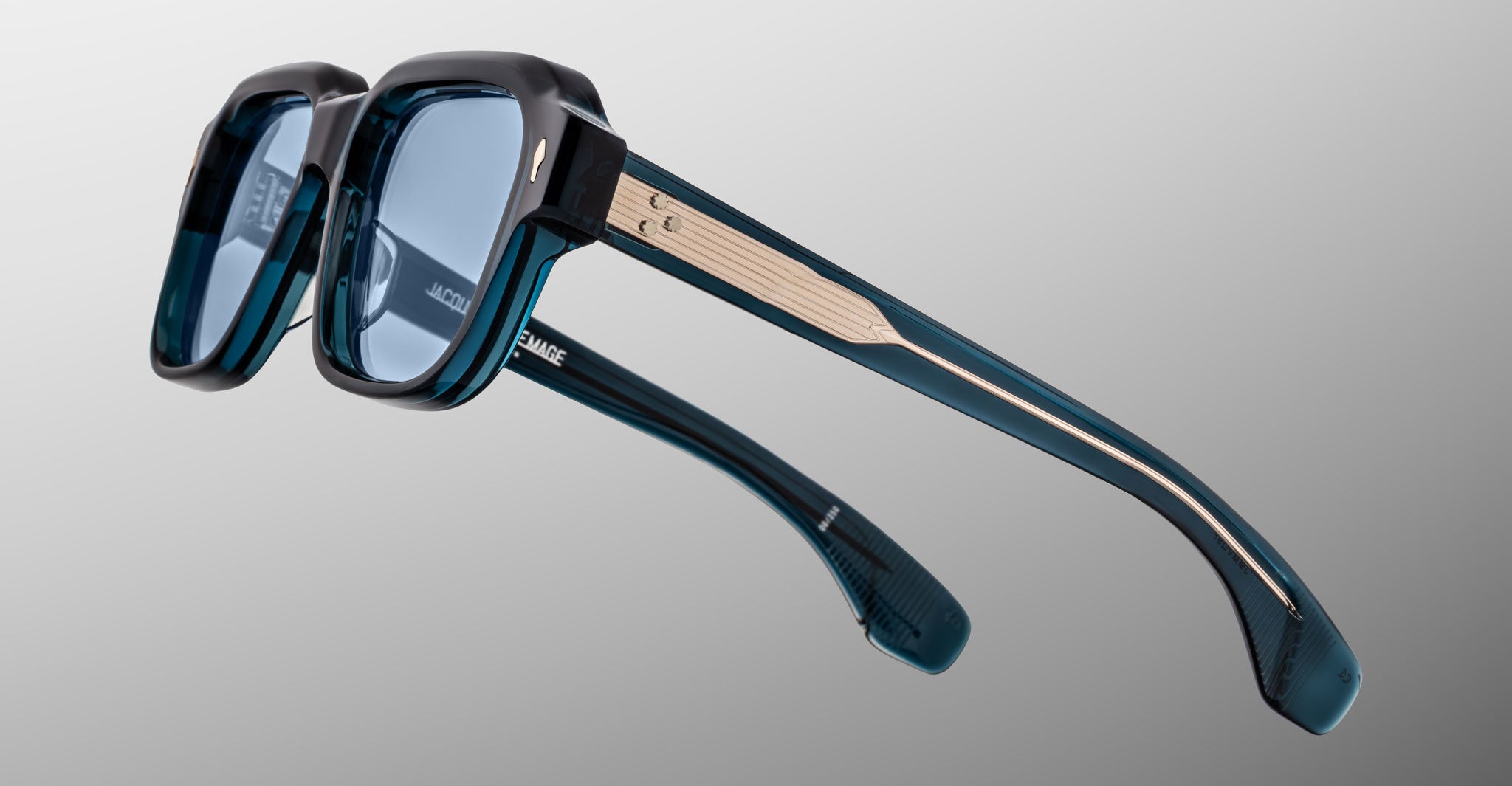 Close-up of Jacques Marie Mage Vivienne sunglasses featuring blue-tinted square lenses, thick teal and black frames, metal arm accents, and a subtle logo inside the temple, set against a gradient gray background.