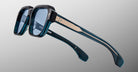 Close-up of Jacques Marie Mage Vivienne sunglasses featuring blue-tinted square lenses, thick teal and black frames, metal arm accents, and a subtle logo inside the temple, set against a gradient gray background.