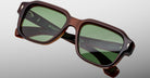 Close-up of Jacques Marie Mage Vivienne brown rectangular sunglasses featuring green-tinted lenses, detailed frame design, and reflective glass, set against a light gray background.