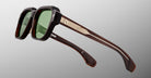 The Jacques Marie Mage Vivienne features stylish rectangular black frames, green lenses, and thick brown arms with metallic accents, displayed on a light gray background.