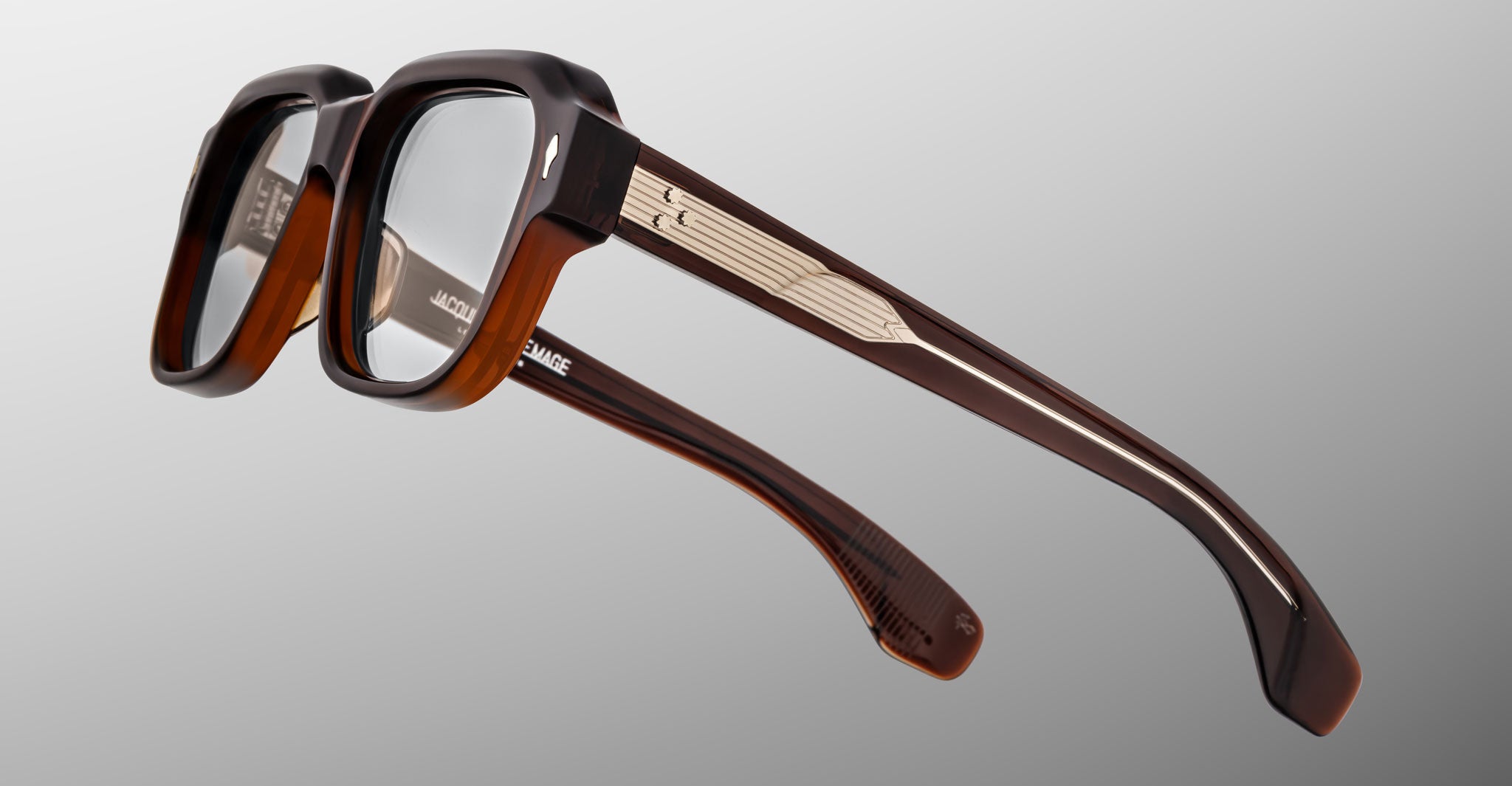 The Jacques Marie Mage Vivienne eyeglasses feature thick dark brown frames, transparent lenses, and detailed metallic temple accents, set against a light gray gradient background.