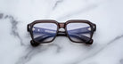 A pair of Jacques Marie Mage Vivienne rectangular sunglasses with dark frames and slightly tinted lenses rests on a white marble surface.