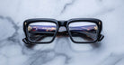 A pair of Jacques Marie Mage Walker thick black rectangular eyeglasses rests on a white marble surface, temples crossed behind the lenses, revealing engraved text inside the left temple.