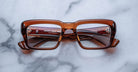 Jacques Marie Mage Walker rectangular, thick-framed brown eyeglasses rest on a white marble surface, shown from the front with visible inner arms and metallic hinge details.