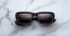 Jacques Marie Mage Walker black square-frame sunglasses with dark lenses rest on a white marble surface, displaying reflections of text and a light source on the lenses.