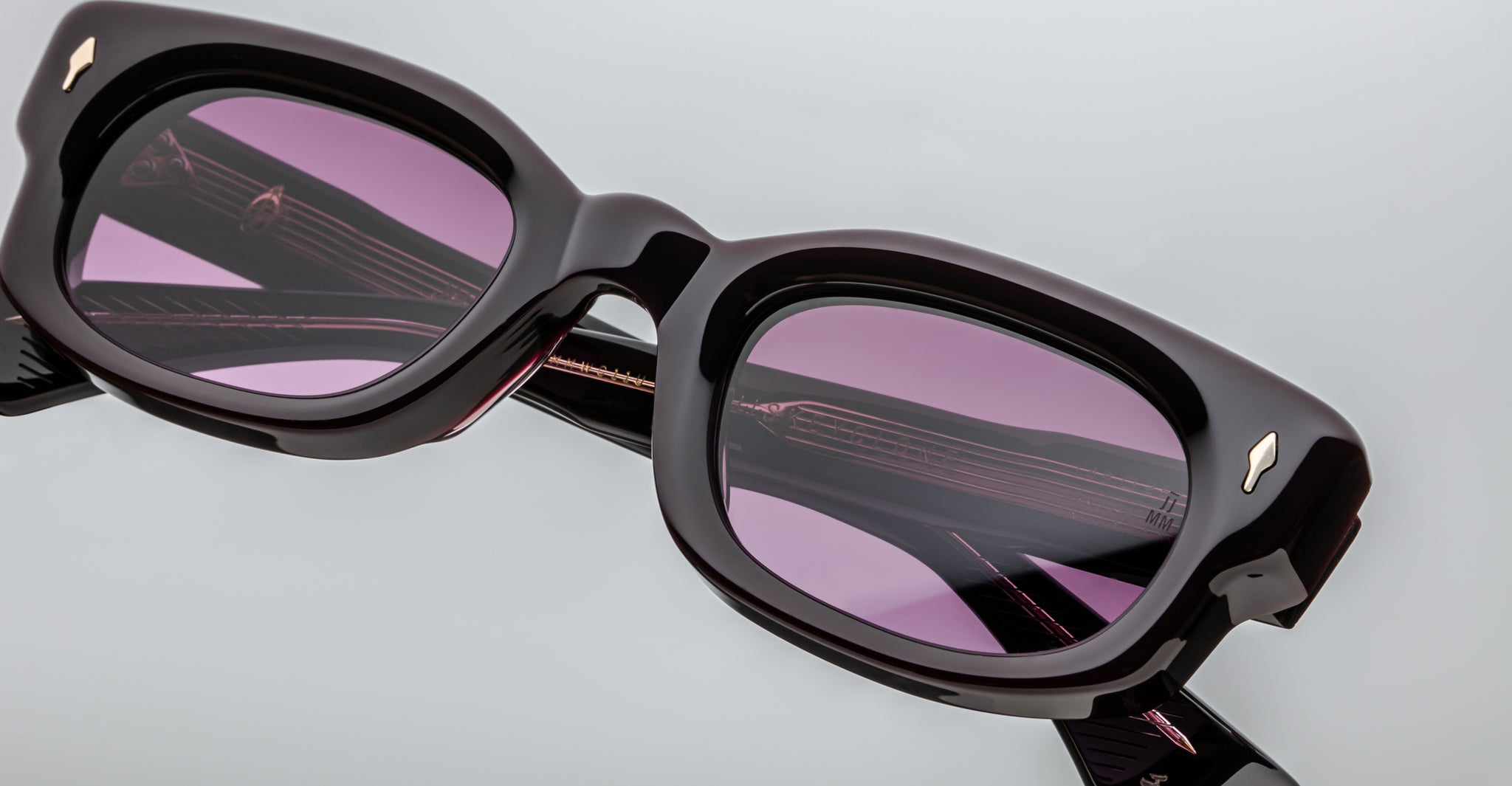 Close-up of Jacques Marie Mage Whiskeyclone black rectangular eyeglasses with purple-tinted lenses, shown against a light gray background. The lenses reflect the glasses’ temples and subtle detailing.