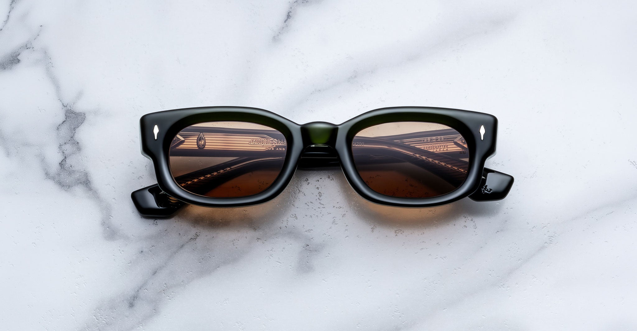 Jacques Marie Mage Whiskeyclone in Borneo sunglasses, dark green acetate frame with translucent olive green accents, warm amber-tinted lenses, bold rectangular silhouette, sculpted bridge, and signature silver-tone arrowhead rivets, shown in a straight-on front view against a white marble background