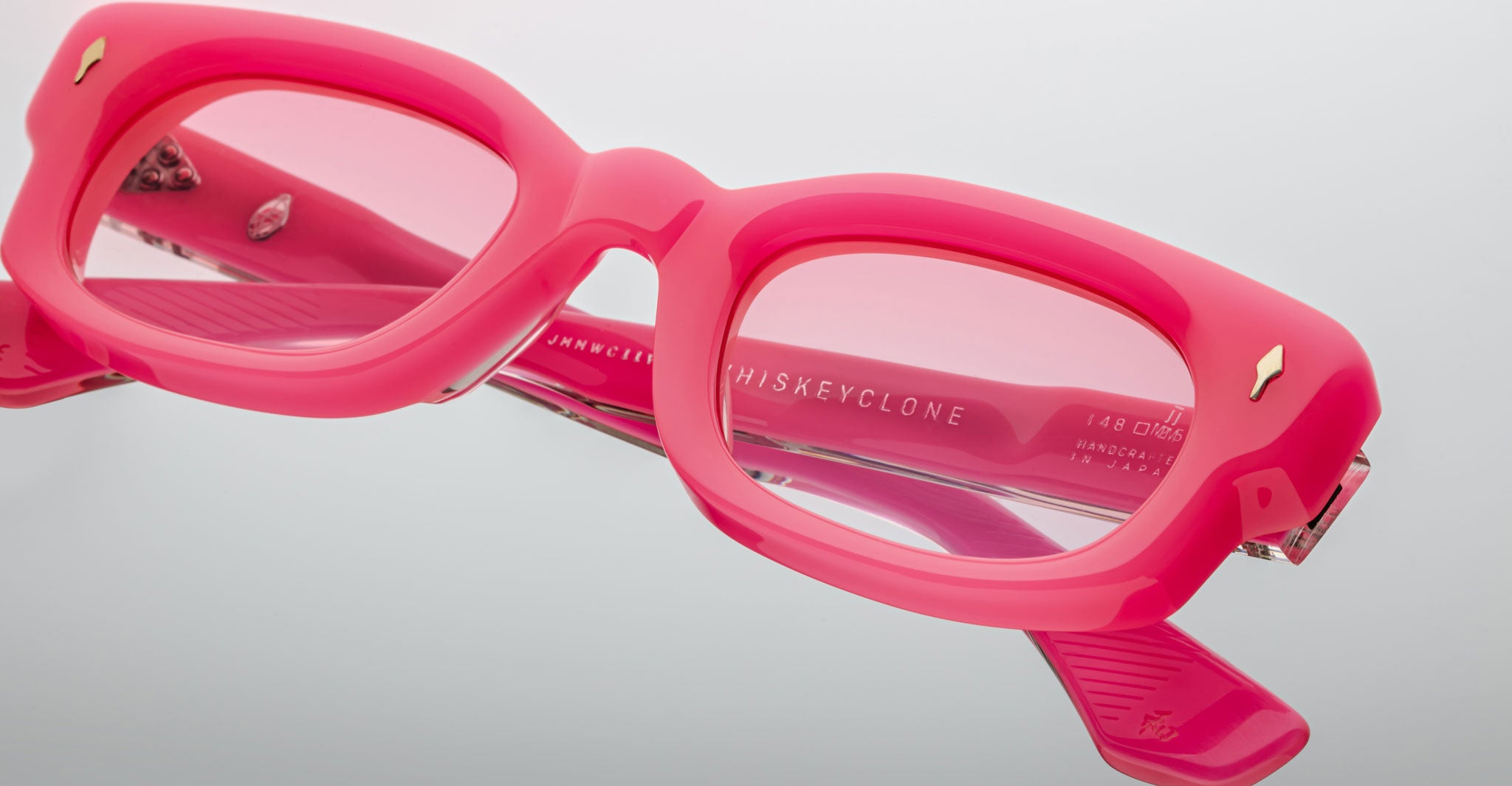 Close-up view of Jacques Marie Mage Whiskeyclone eyeglasses—thick, rectangular frames in bright pink with pink-tinted lenses—shown on a white background. Brand and model details are visible inside the frame.