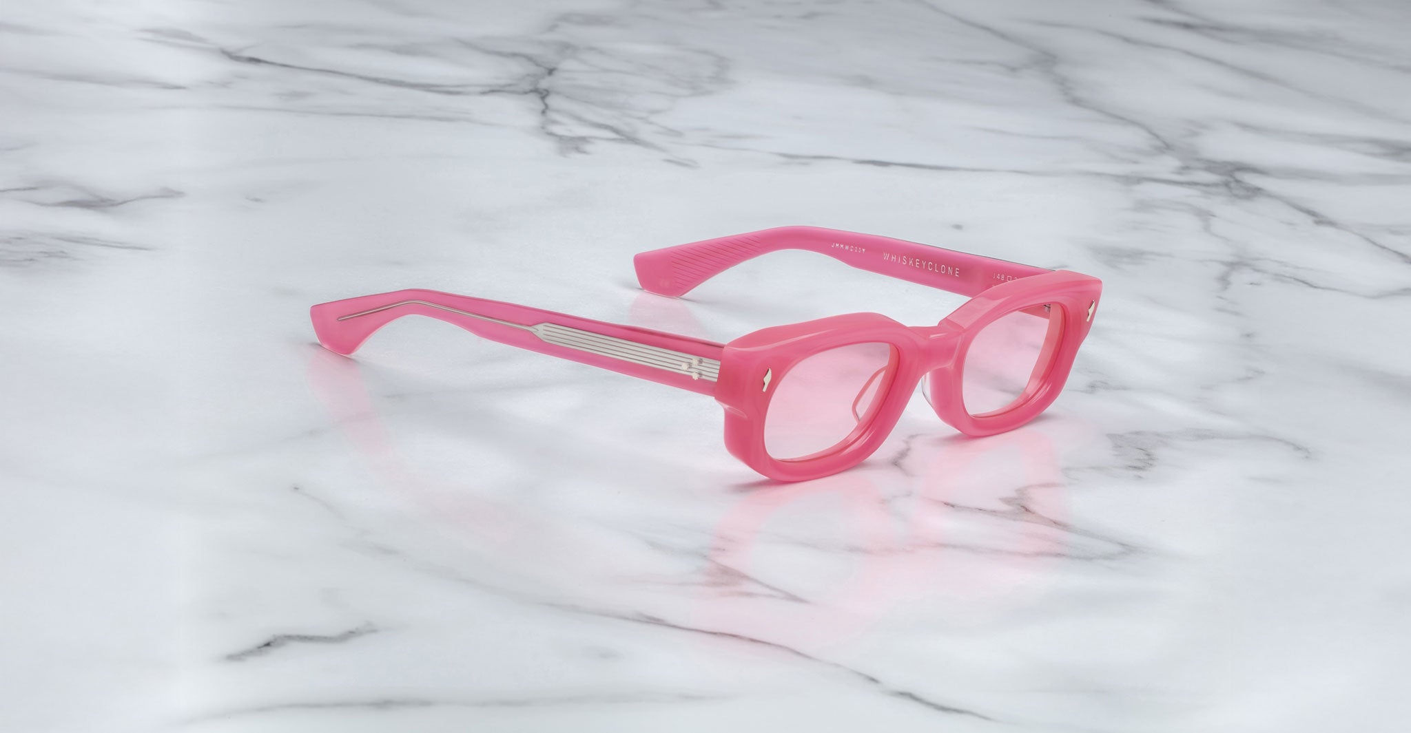 Jacques Marie Mage Whiskeyclone eyeglasses with pink thick rims and arms rest on a white marble surface with gray veins. The arms feature silver horizontal detailing near the hinges.