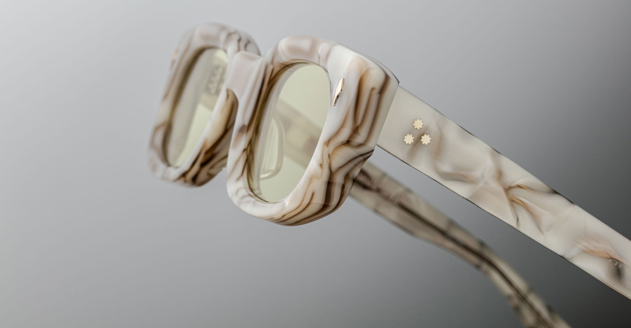 A close-up of Jacques Marie Mage Whiskeyclone square-framed sunglasses featuring a marble pattern, light-tinted lenses, and small gold star-shaped temple details set against a neutral background.