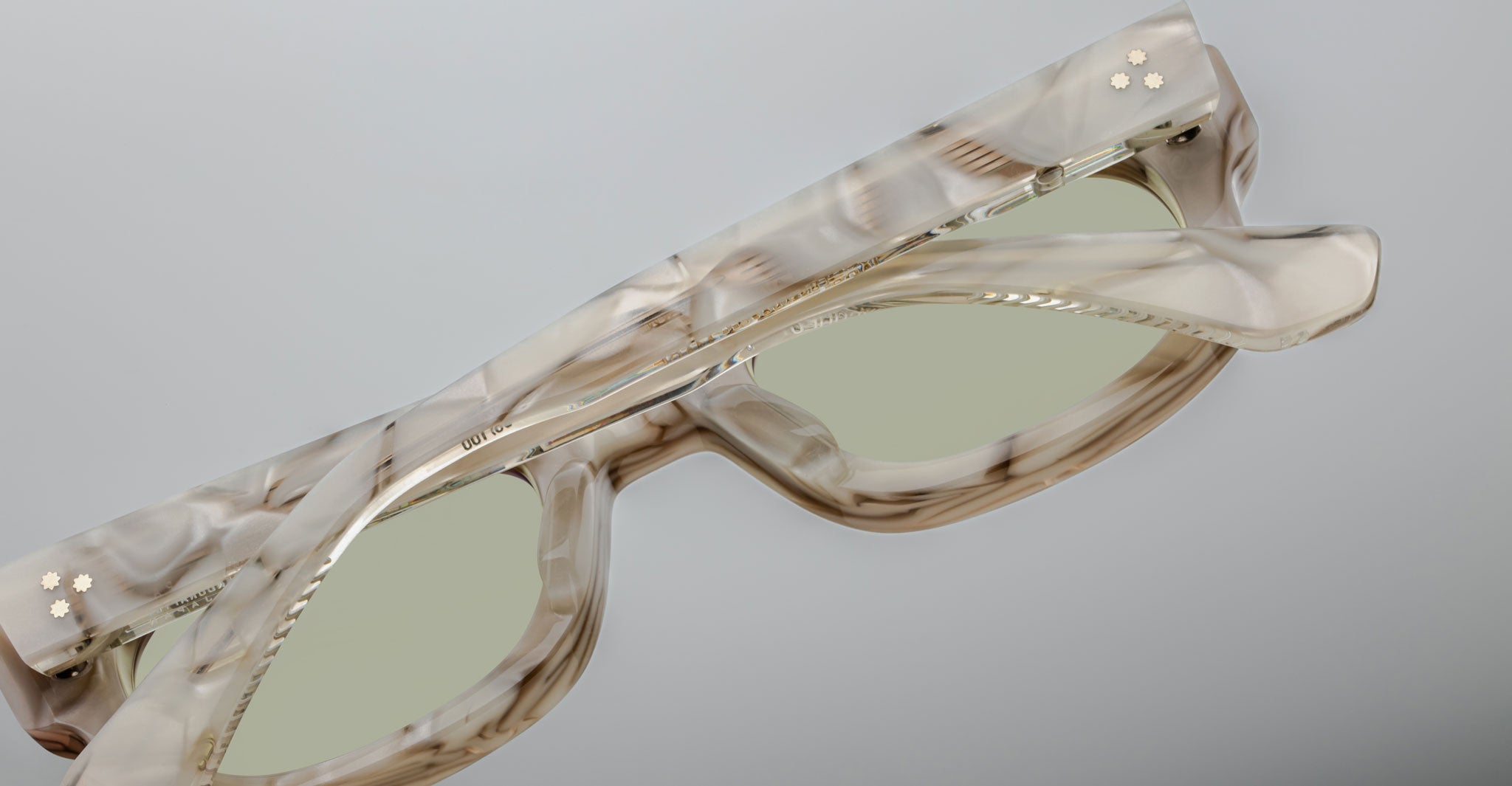 A close-up of Jacques Marie Mage Whiskeyclone rectangular sunglasses with a marbled beige and brown frame, light green lenses, and two small gold flower-shaped studs on one arm, set against a plain gray background.