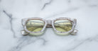 A pair of Jacques Marie Mage Whiskeyclone rectangular eyeglasses with a marble pattern and yellow-tinted lenses rests on a white marble surface.