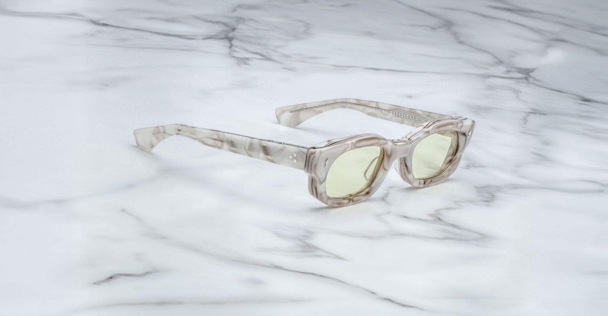 The Jacques Marie Mage Whiskeyclone sunglasses feature a clear marble-patterned frame and arms with a translucent, milky look, paired with light yellow tinted lenses, and are displayed on a white marble surface.