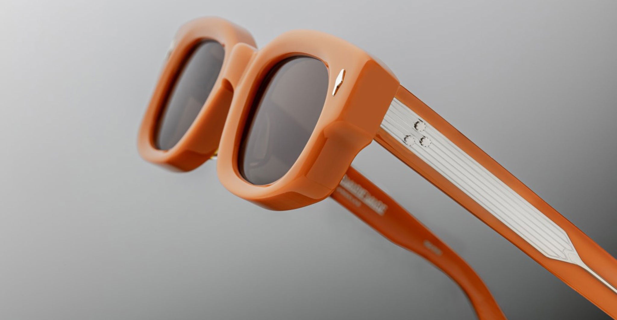 Close-up view of Jacques Marie Mage Whiskeyclone orange rectangular sunglasses with dark lenses and metallic accents on the arms, set against a plain light gray background.