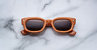 The Jacques Marie Mage Whiskeyclone sunglasses feature square-shaped, thick light brown frames with dark lenses, displayed on a white marble surface with gray veining.