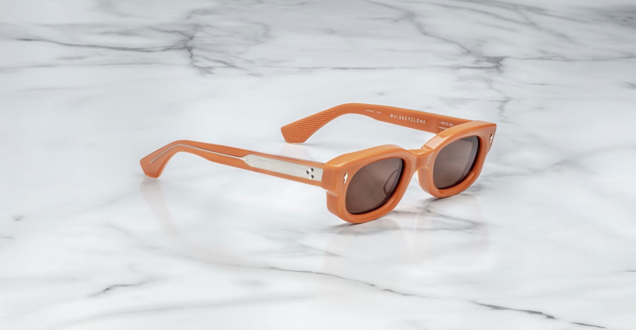A pair of Jacques Marie Mage Whiskeyclone orange rectangular sunglasses with dark lenses and silver arm detailing, placed on a white marble surface.