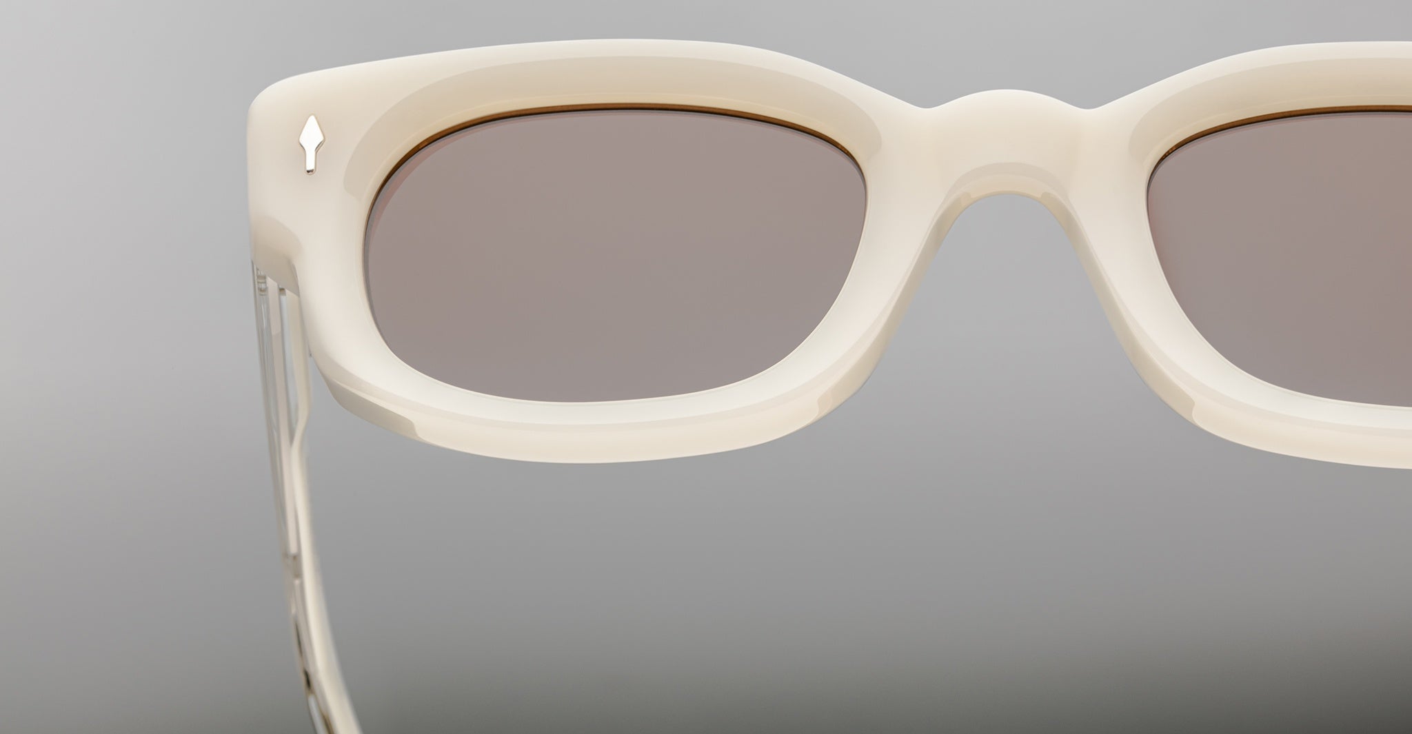 Close-up view of Jacques Marie Mage Whiskeyclone sunglasses featuring thick off-white rims, light brown lenses, and a metallic arrow detail on the temple, set against a neutral gray background.